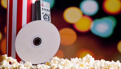 Closeup red white striped container standing up with popcorn lying around, dvd disc and remote control leaning on box, low angle, flashy vivid lights background