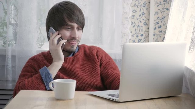 Happy Young Man Freelancer Working with Modern Laptop and Phone in Cafe taking Cup of Coffee