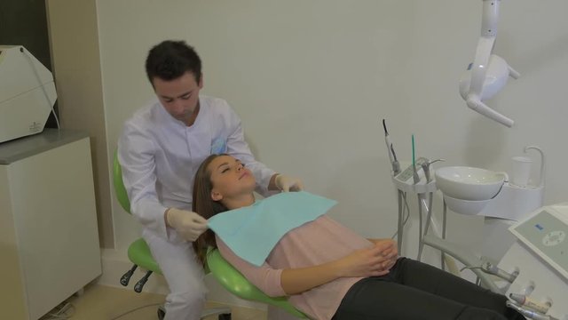 Male Dentist Fixing a Paper Napkin on Chest Put the Mask on and Starts to Examine a Teeth Client Young Woman is Sitting in a Dentist Chair Dental Clinic