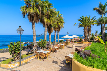 Palm trees and restaurant tables on coastal promenade in Puerto de la Cruz town, Tenerife, Canary Islands, Spain