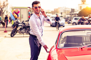 Young handsome man with phone opening red car door - Attractive businessman talking at phone is getting into sport vehicle - Successful male person using mobile technology outdoors - Lifestyle concept