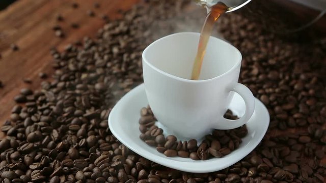 Pouring flavored coffee from coffee pot in white cup surrounded by coffee beans on wooden table. Close up view
