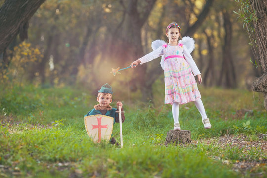 Cute little girl dressed up as a fairy playing with a boy dressed up as a knight sitting in a magical forest