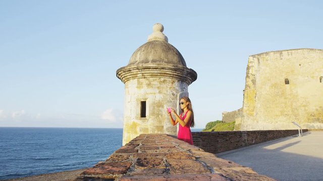 Puerto Rico travel tourist woman in San Juan, taking photo with phone looking at the fort Castillo San Felipe Del Morro, famous Caribbean cruise ship destination of Old San Juan, Puerto Rico, USA.