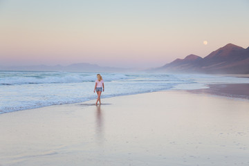 Woman walking on sandy beach in sunset. Waves sweeping away her traces in sand. Beach, travel,...