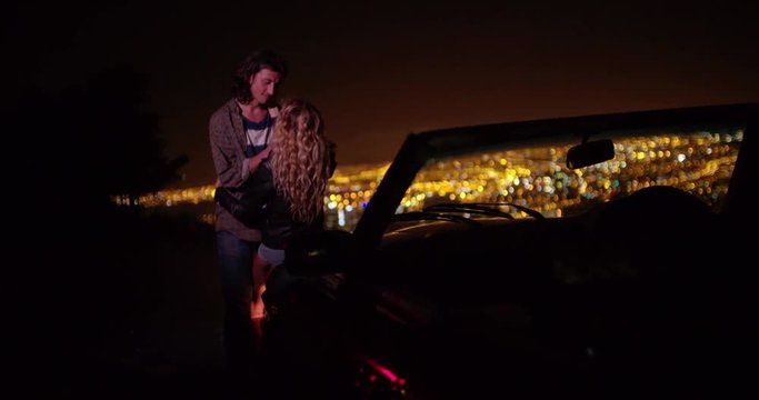 Young couple hugging and looking at night city lightsnext to a convertible