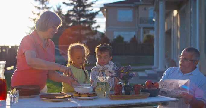 Cute grandkids cooking a cake together with grandmother to help her with setting outdoor dining table while their grandfather reading newspaper