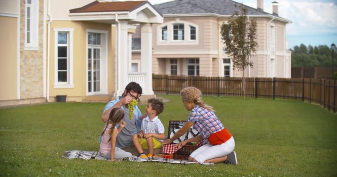 Young Mother, Father And Their Little Son And Daughter Enjoying Their Picnic In The Backyard: Mom Taking Food From Basket And Dad Giving Grape To Kids