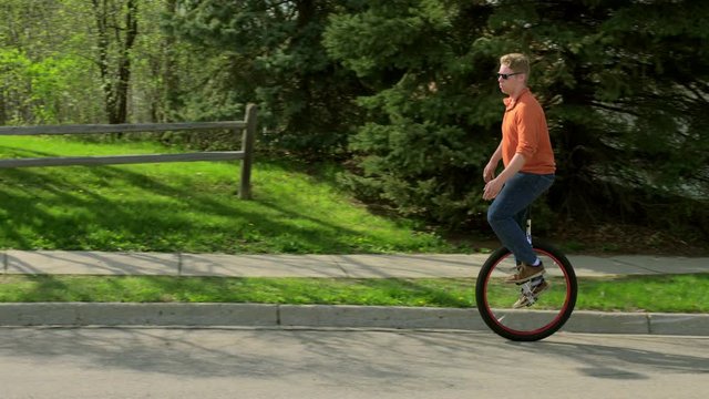 Young man in sunglasses riding a unicycle on a Mid Western suburban street.   Wide shot recorded in slow motion 4K at 60fps on a sunny spring day.