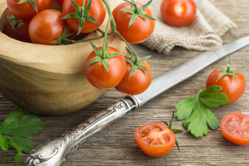 Heap of cherry tomatoes in bowl, with knife and some cut in half, on wooden surface.