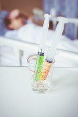 Syringes in a glass measuring cup with blurred illness boy lying on sickbed.
