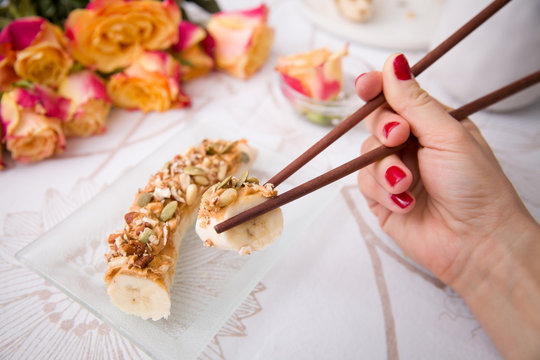 Banana sushi servered with nuts and granola on a glass plate. Hands with Japanese sticks. Woman eating healthy snack. Flowers and fruit breakfast or dessert.