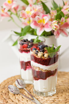 Homemade granola and parfait serverd in high glasses with yogurt and organic raspberries, nuts and mint. Delicious snack for healthy nutrition.  Breakfast on a straw tablemat with spoons and flowers