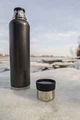 Obraz premium A man takes a hand in mitten cup of hot tea from a thermos, which stands on an ice floe on the river a sunny day