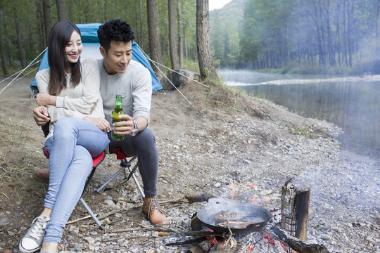 Young couple sitting beside campfire preparing food