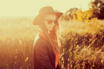 closeup attractive beautiful brunette woman with fluffy long hairs posing standing in a Park, autumn meadow, sunset light, in a black hat, a denim vest and shorts,spring mood, sadness,outdoor portrait