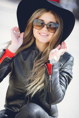 Portrait of a young woman with a beautiful smile,blonde long hair,dressed in a black leather jacket, black hat with large brim and black leather pants,wears dark sun glasses, sitting alone outside