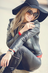 Portrait of a young woman with a beautiful smile,blonde long hair,dressed in a black leather jacket, black hat with large brim and black leather pants,wears dark sun glasses, sitting alone outside