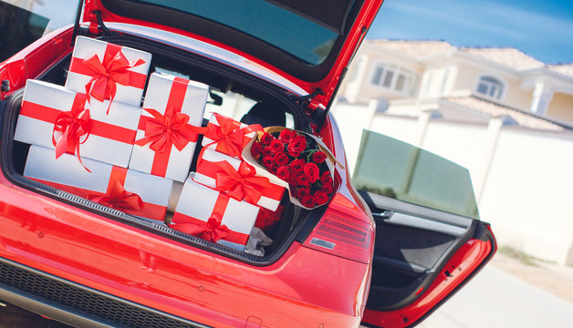 White boxes with gifts tied with red ribbons with large red bows and a huge bouquet of red roses,neatly stacked in the open trunk of the big red car,the concept of celebration and love