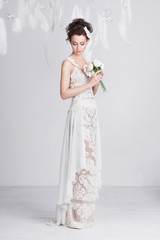 Tall and slim young bride in a luxurious lace wedding dress. She has a beautiful tanned skin. The dress is translucent. White feathers around her. She is holding a bouquet of white and pink roses.