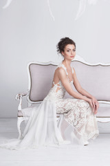 Beautiful young bride in a luxurious lace wedding dress on a white vintage sofa. She is an elegant and slim, she has tannes skin, dark hair and gray eyes. The dress has a translusent silk train.