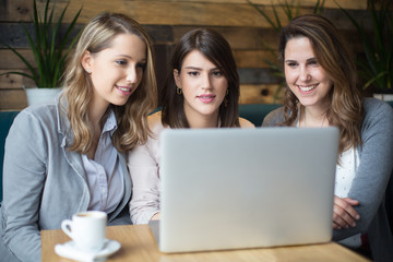 Three young women sitting at cafe drinking coffee and looking at laptop
