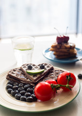 On the table there is a plate filled with chocolate cakes, blueberries, tomato snacks, fruit, a glass of lemonade