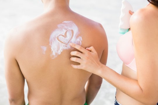Woman making a heart symbol on mans back while applying a sunscreen lotion at beach