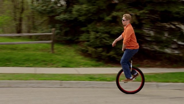 Young man in sunglasses riding a unicycle around a street in a suburban neighborhood.  Rides out of frame at end.  Wide shot, recorded in 4K on a spring day in the suburban Mid West.