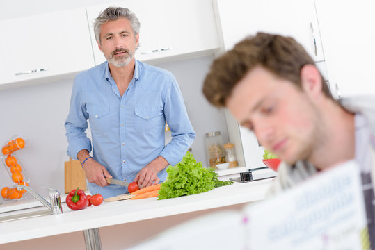 Man preparing vegetables, looking forward at younger man's book