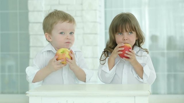 Little boy and girl eat red apples.We look at each other and then leave. Children in white shirts next to the table in a bright room. Against the background of light brick wall and the window.