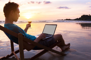 young business man with cocktail working on laptop on the beach at sunset, freelance job online, focus on the screen