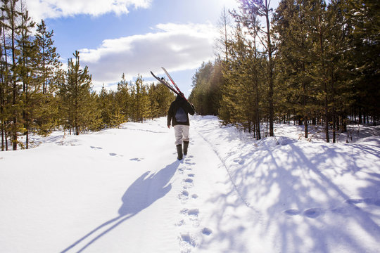 Rear view of a man carrying skis while walking on snowy field