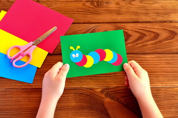 Children hands holding paper applique caterpillar. Paper sheets, scissors on a brown wooden background