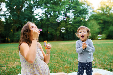 Young pregnant mother resting with her son in summer park and bl