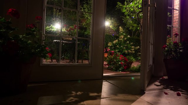 Dark room with an open French door leading to the illuminated patio full of colorful flowers and plants in the night. Dolly shot.