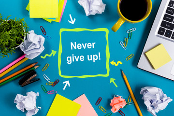 Never give up. Office table desk with supplies, white blank note pad, cup, pen, pc, crumpled paper, flower on blue background. Top view