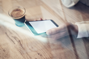 Closeup photo of modern smartphone holding in female hands. Blank screen ready for business information. Cup espresso on the table, horizontal, visual effects. 