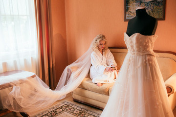 Beautiful blonde bride in robe posing near white wedding dress