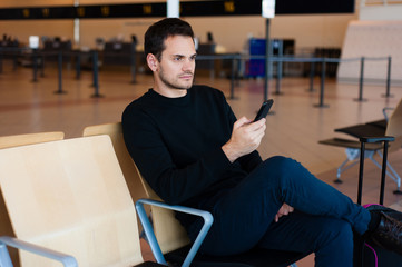 Handsome man in casual wear sitting in the hall of the airport terminal with his mobile phone while while waiting for his flight with hand luggage suitcase.