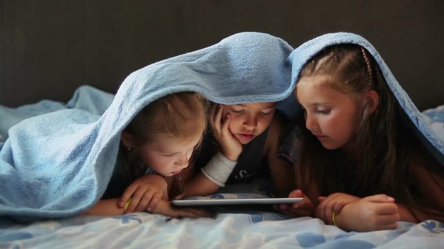 Children play on the tablet while lying under a blanket