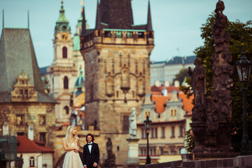 Fototapeta premium Beautiful blonde bride & handsome groom posing on Prague bridge,