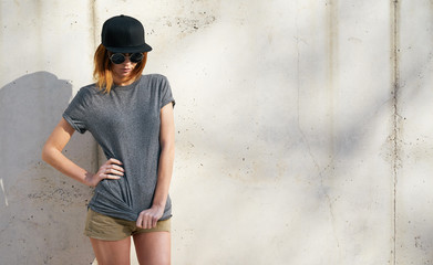 beautiful young woman wearing in a gray blank t-shirt and black blank cap posing against a background of a concrete wall in the rays of the setting sun