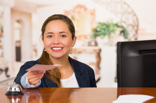 Young brunette hotel receptionist with friendly smile handing over key to client across desk, customers point of view
