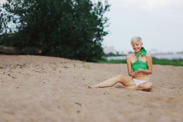 Happy young blond woman in a swimsuit sits on the sand on a beach and laughing