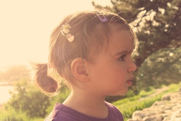 Portrait of a little dark haired girl with pony tail, on a sunny summer day, in a park. Image filtered in faded, washed out, retro, vintage, Instagram style with lens flare.