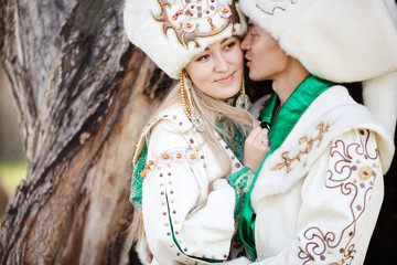 Couple in ethnic costumes embrace on background of textured wood, groom kisses bride at cheek.