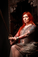 Studio shot of beautiful young redhead girl with multiple tattoo in chainded armour with the sword
