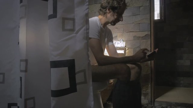 Dolly reveal shot of a man sitting on the toilet with his cell phone