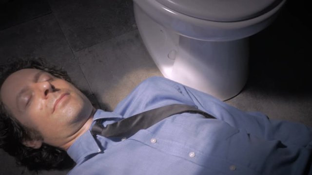 Man in a shirt and tie answers his phone while laying next to a toilet
